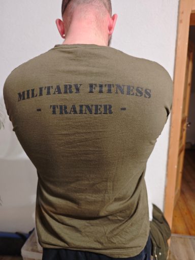 Mann mit militärischem Fitness-Trainer-Shirt, Rückenansicht, in einem Innenraum.